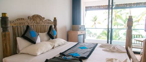 A hotel room with a large bed, a wooden headboard, a small bedside table, and a view of palm trees through the window.