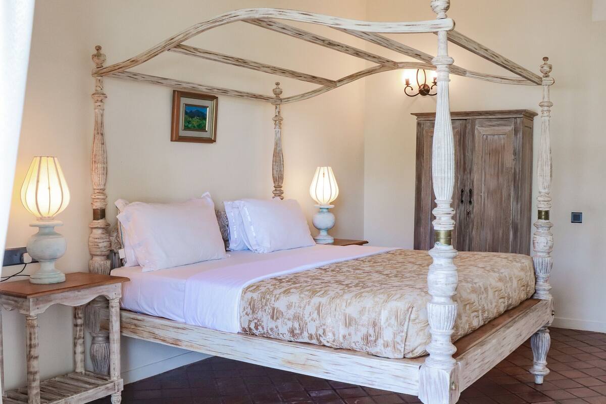 A four-poster bed with white bedding, two bedside lamps, and a wooden wardrobe in the background.