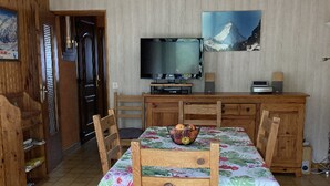 Un coin repas avec des chaises en bois et une table recouverte d’une nappe à motifs floraux. Un téléviseur est fixé au mur, au-dessus de la table.
