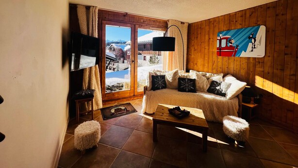 Una sala de estar acogedora con un sofá, una mesa de centro y un reposapiés mullido. Un televisor está montado en la pared, y una ventana muestra vistas de un paisaje nevado.