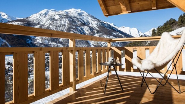 Eine Holzterrasse mit einem Stuhl und einem kleinen Tisch, mit Blick auf schneebedeckte Berge.