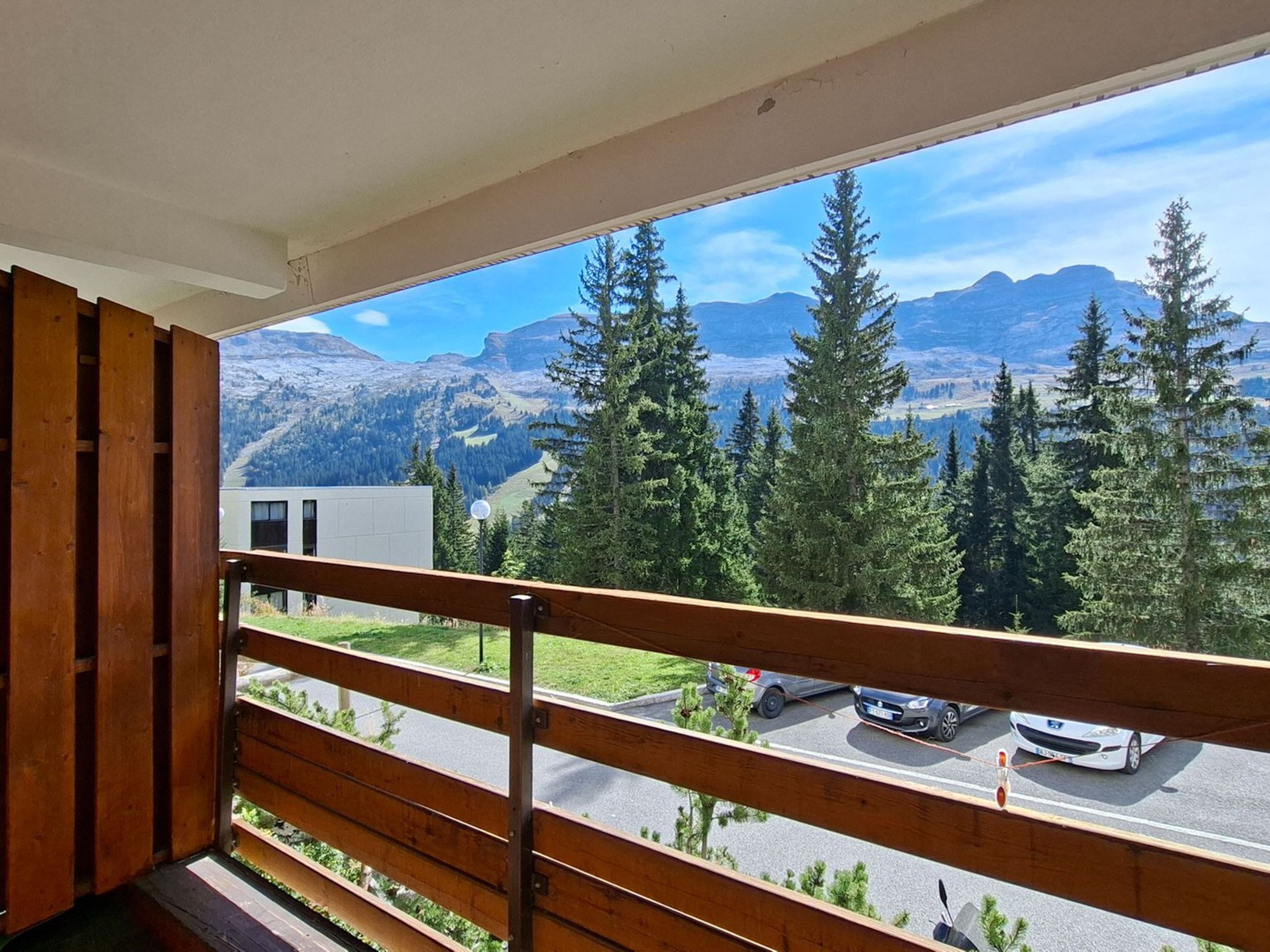 Un balcón con vista a un paisaje montañoso, un estacionamiento con coches y un edificio al fondo.