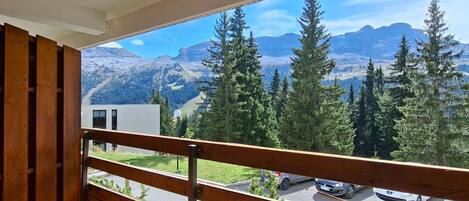 A balcony with a view of a mountainous landscape, a parking area with cars, and a building in the background.