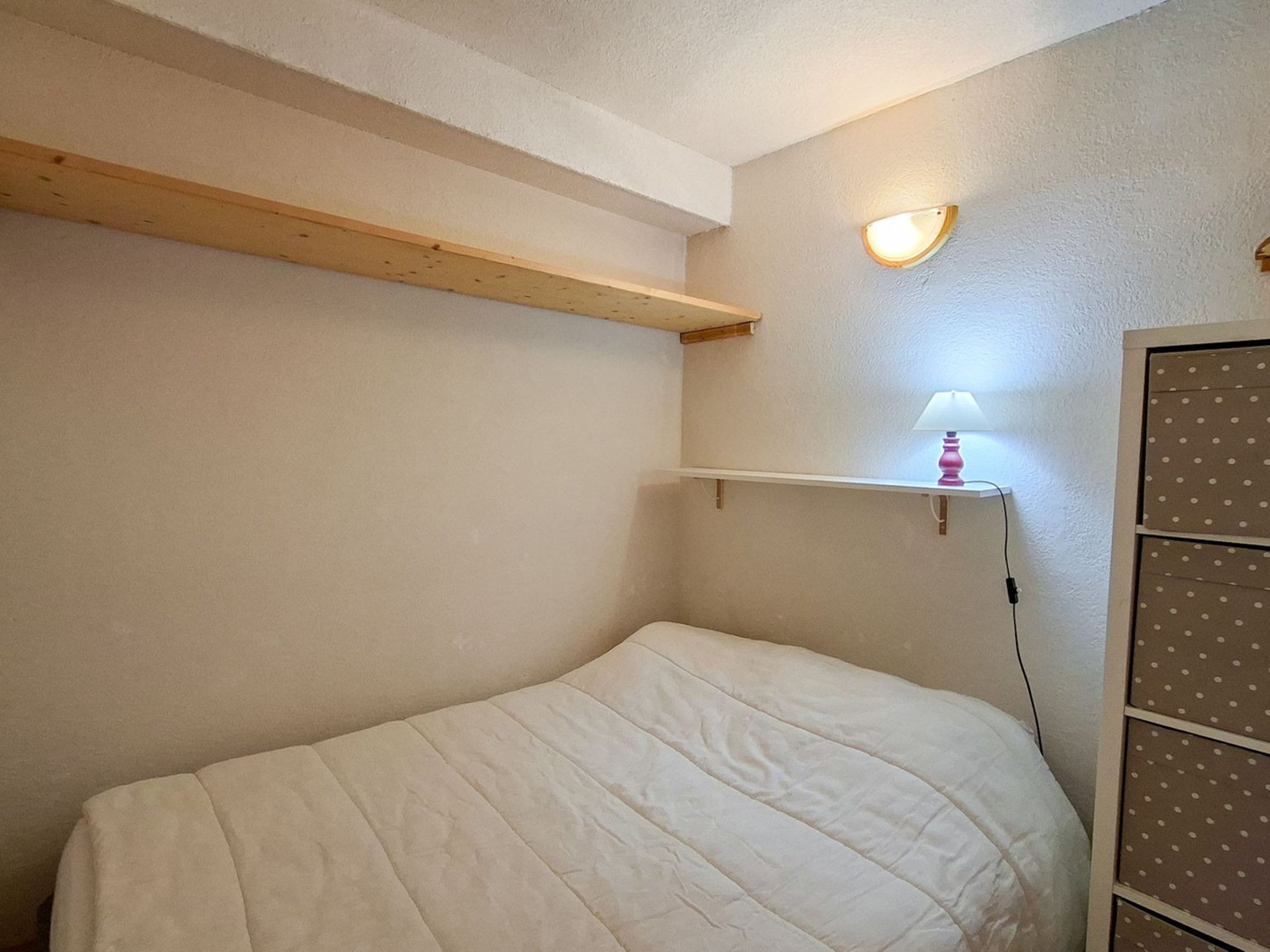 A bedroom with a single bed, a shelf with a lamp, and a wall-mounted light fixture.