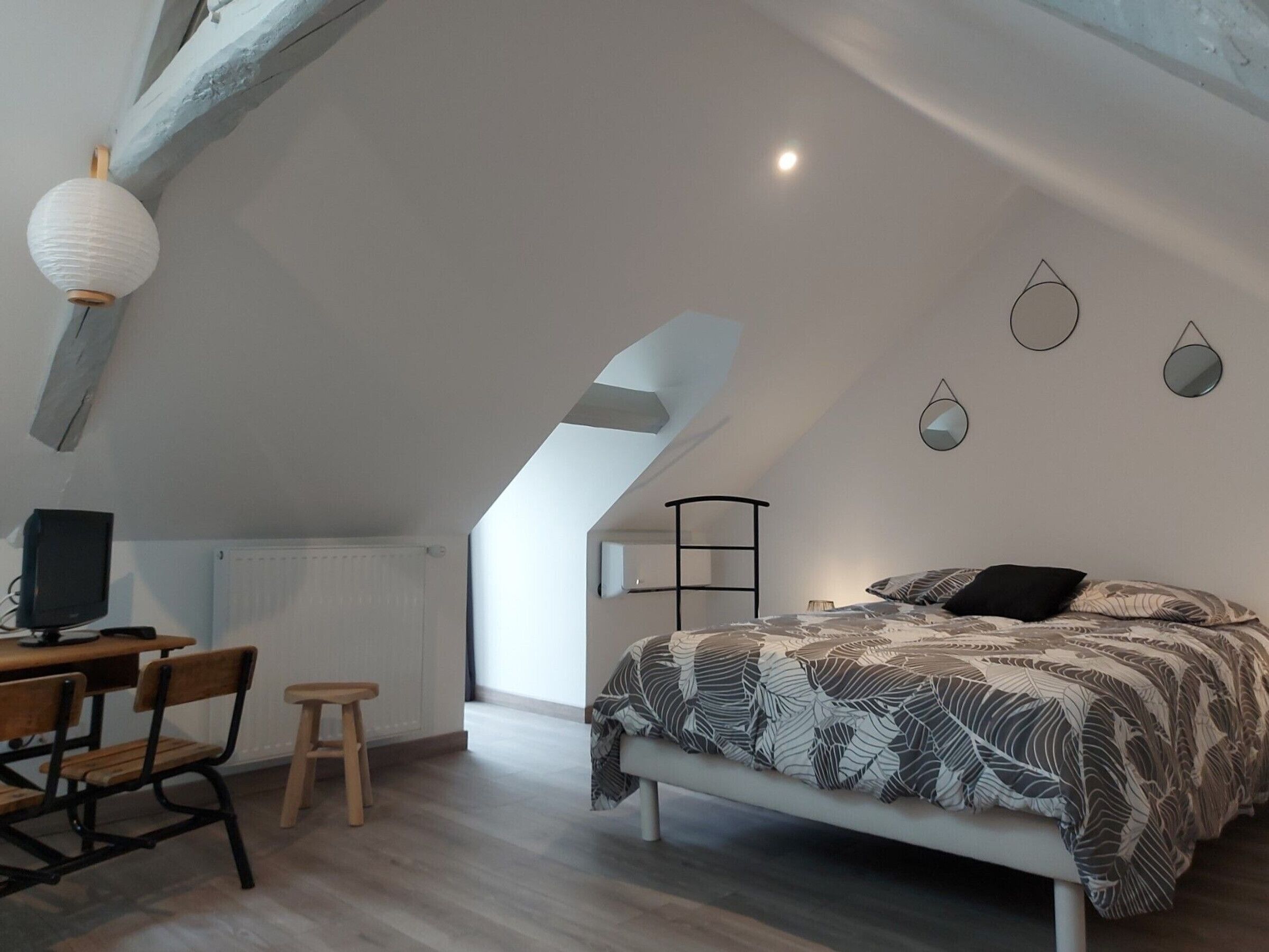 A bedroom with a sloped ceiling, a bed with patterned bedding, a desk with a chair, and a television.