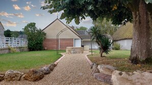 Une maison avec une allée en gravier menant à un patio en briques, entourée d’une pelouse verdoyante et d’un grand arbre.
