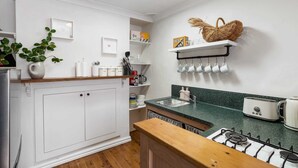 Cocina con armarios blancos, estufa a gas, encimera verde y una estantería con tazas.