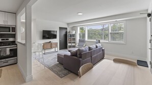 A modern living room with a grey sectional sofa, a flat-screen TV, a dining area with white chairs, and a kitchen with built-in appliances. - West Taylor Run Hotel (Alexandria)