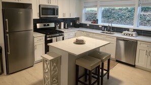 A modern kitchen with white cabinets, stainless steel appliances, a central island with stools, and a window with a view of greenery. - West Taylor Run Hotel (Alexandria)