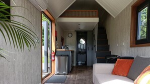 A compact living space with a sofa, a colorful rug, a kitchen area, and a staircase leading to an upper level. - The Tiny House (Newton Abbot)