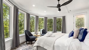 A bedroom with a large bed, two chairs, a ceiling fan, and multiple windows offering a view of trees.