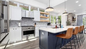 A modern kitchen with white cabinets, a central island with bar stools, a built-in oven, and a stone fireplace.