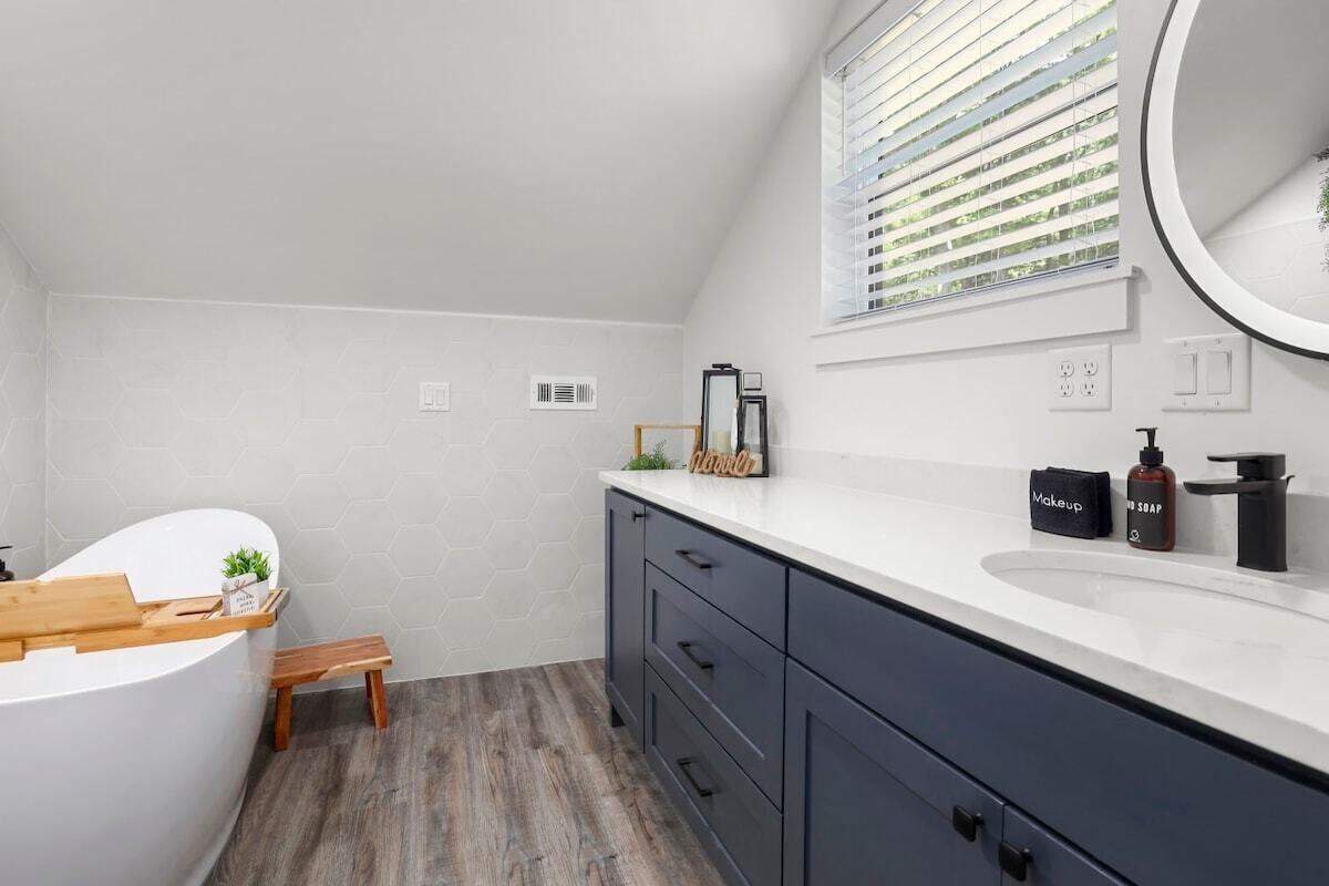 A modern bathroom with a white bathtub, dark cabinets, a wooden bench, and a large mirror above the sink.