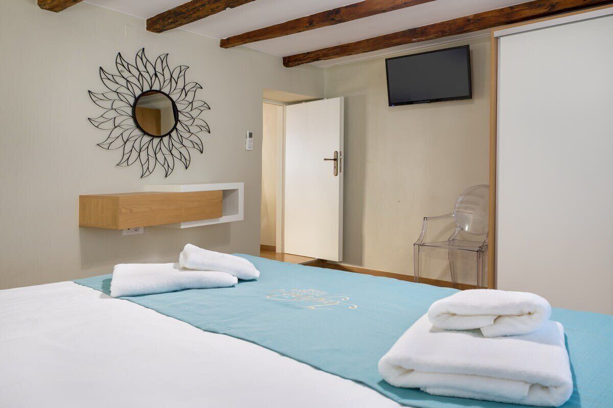Une chambre d’hôtel avec un lit, une couvre-lit bleue, une étagère en bois avec un miroir, un téléviseur fixé au mur et une porte donnant sur une autre pièce.