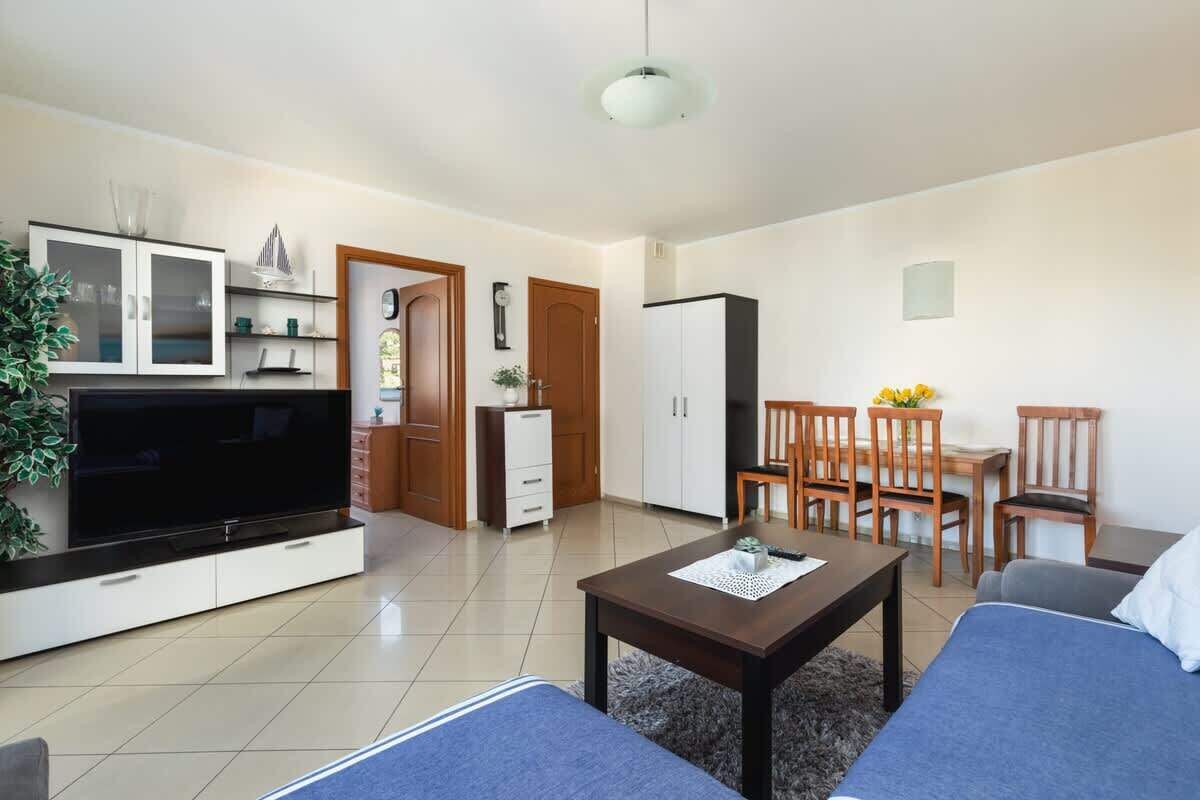 Uma sala de estar moderna com uma TV de tela plana, uma mesa de centro e um sofá. Há uma área de jantar com cadeiras de madeira e uma pequena mesa. O cômodo tem uma paleta de cores neutras e uma janela grande.