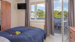 Une chambre avec un lit, une télévision, un balcon avec une table et des chaises, et une vue sur les arbres et les bâtiments.