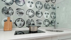 Une cuisine avec un plan de travail blanc, une planche à découper en bois, une casserole sur une plaque de cuisson à induction et un évier. Le mur derrière le plan de travail est décoré d’une série de portraits circulaires.