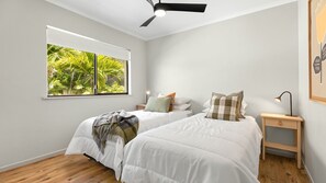 Two single beds with white duvets and multiple pillows, a large window with a view of lush greenery, and a ceiling fan.