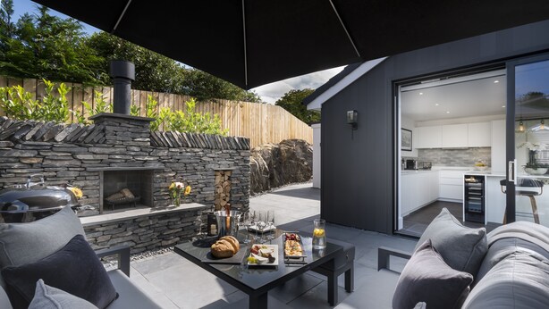 Un patio al aire libre con chimenea de piedra, una mesa de comedor con comida y una cocina moderna visible a través de puertas de vidrio.