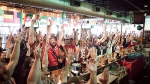 A crowded bar with raised hands, various flags, and a bar counter with bottles and glasses. - Aquarius Rising (Atlanta)