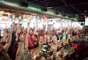 A crowded bar with raised hands, various flags, and a bar counter with bottles and glasses. - Aquarius Rising (Atlanta)