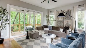 A spacious living room with a large sofa, a coffee table, and a ceiling fan. There is a sliding glass door leading to a balcony with a wooden deck. - Aquarius Rising (Atlanta)