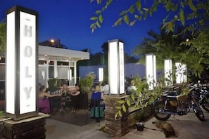 Entrada de hotel con un letrero que dice "HOLY" y un área de estar al aire libre cubierta donde personas están cenando.