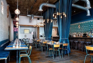 Un restaurante con mesas largas compartidas, cortinas azules y una zona de bar con taburetes.
