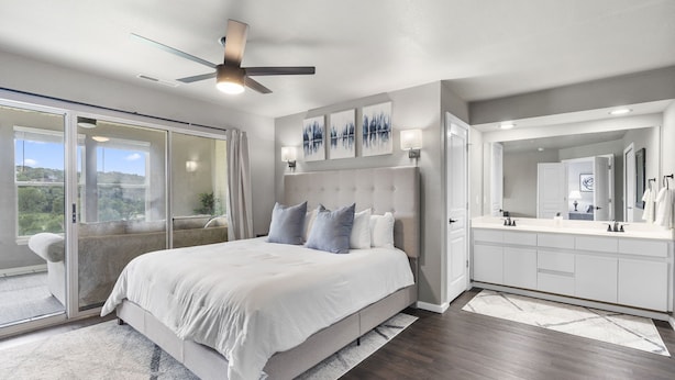 A modern bedroom with a large bed, a ceiling fan, a sliding glass door, a bathroom with a vanity and mirror, and artwork on the walls. - Modern 2-Bed Condo with Golf Course Views in Branson (Branson)
