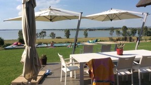 Un patio al aire libre con mesas y sillas blancas, dos sombrillas grandes y vistas a un lago y una piscina al fondo.