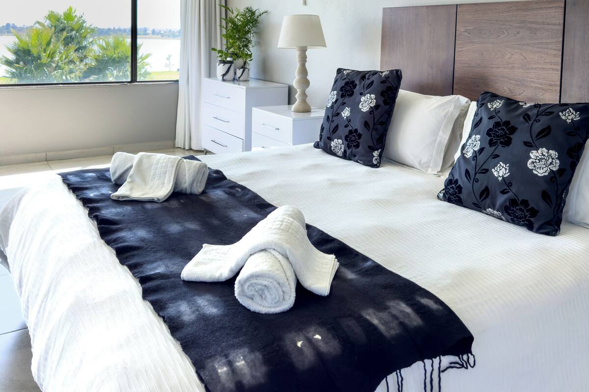 A modern hotel room with a large bed, a nightstand, a lamp, and a view of the outdoors.