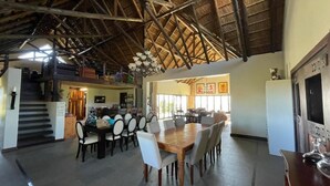 Un comedor espacioso con techo alto y vigas de madera, una mesa central rodeada de sillas, y una zona de cocina al fondo.