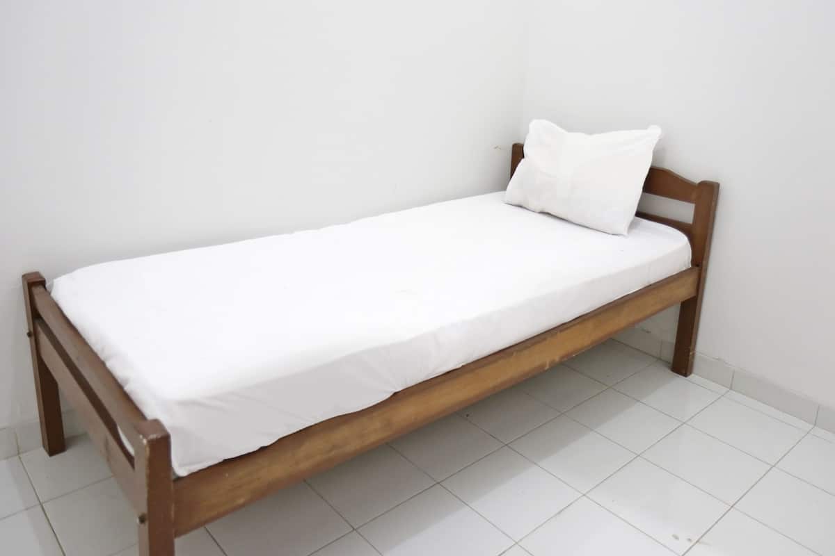 Un simple lit en bois, avec un matelas et un oreiller blancs, dans une pièce dont le sol et les murs sont recouverts de carreaux blancs.