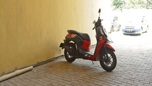 A red scooter parked against a yellow wall in a covered parking area with a brick floor. - OYO Life 92925 Kost Bu Mala 2 Syariah (Sukorejo)