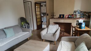 A living room with a grey sofa, a white armchair, a wooden coffee table, and a white cabinet. There is a bookshelf with books and a small table with a chair. A baby stroller is visible near the door.