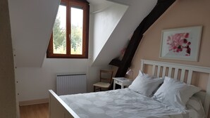 A bedroom with a sloped ceiling, a bed with white bedding, a chair, a desk, a radiator, and a window with a view of greenery.