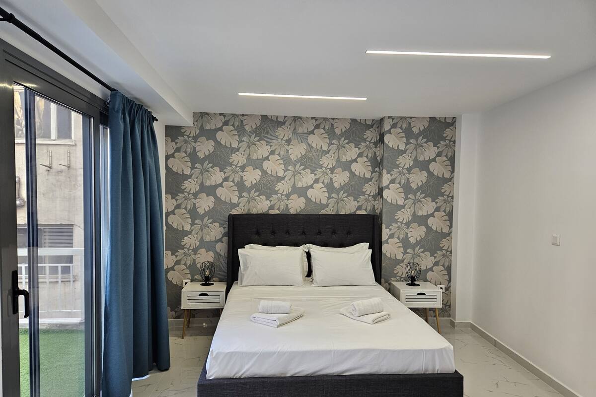 Um quarto moderno com uma cama grande, duas mesas de cabeceira, uma janela com cortinas e um papel de parede floral.
