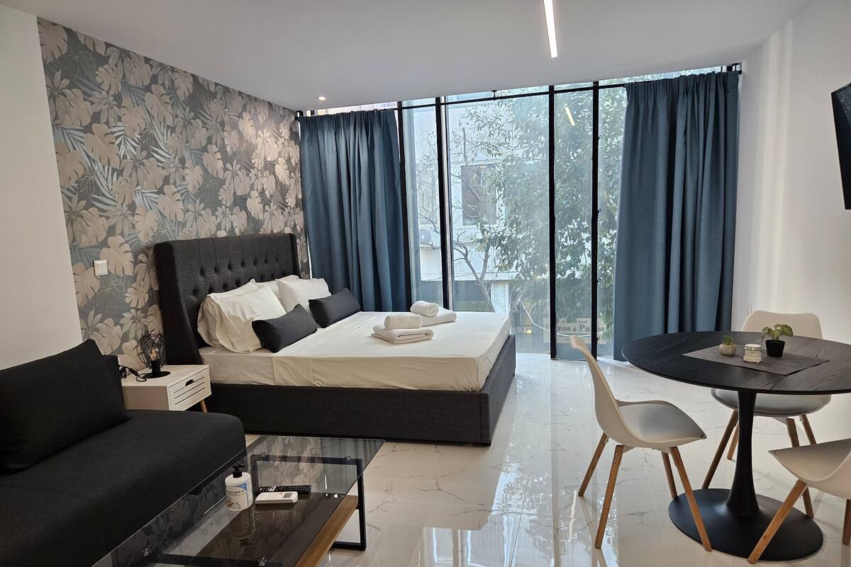 Quarto de hotel moderno com uma cama grande, um sofá, uma mesa de centro de vidro, uma mesa de jantar com cadeiras e vista para a cidade por grandes janelas.