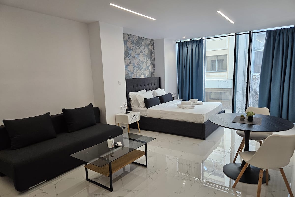 Quarto de hotel moderno com sofá preto, mesa de centro de vidro, cama com roupa de cama branca, pequena mesa lateral, mesa de jantar redonda com uma cadeira e janelas grandes com cortinas azuis.