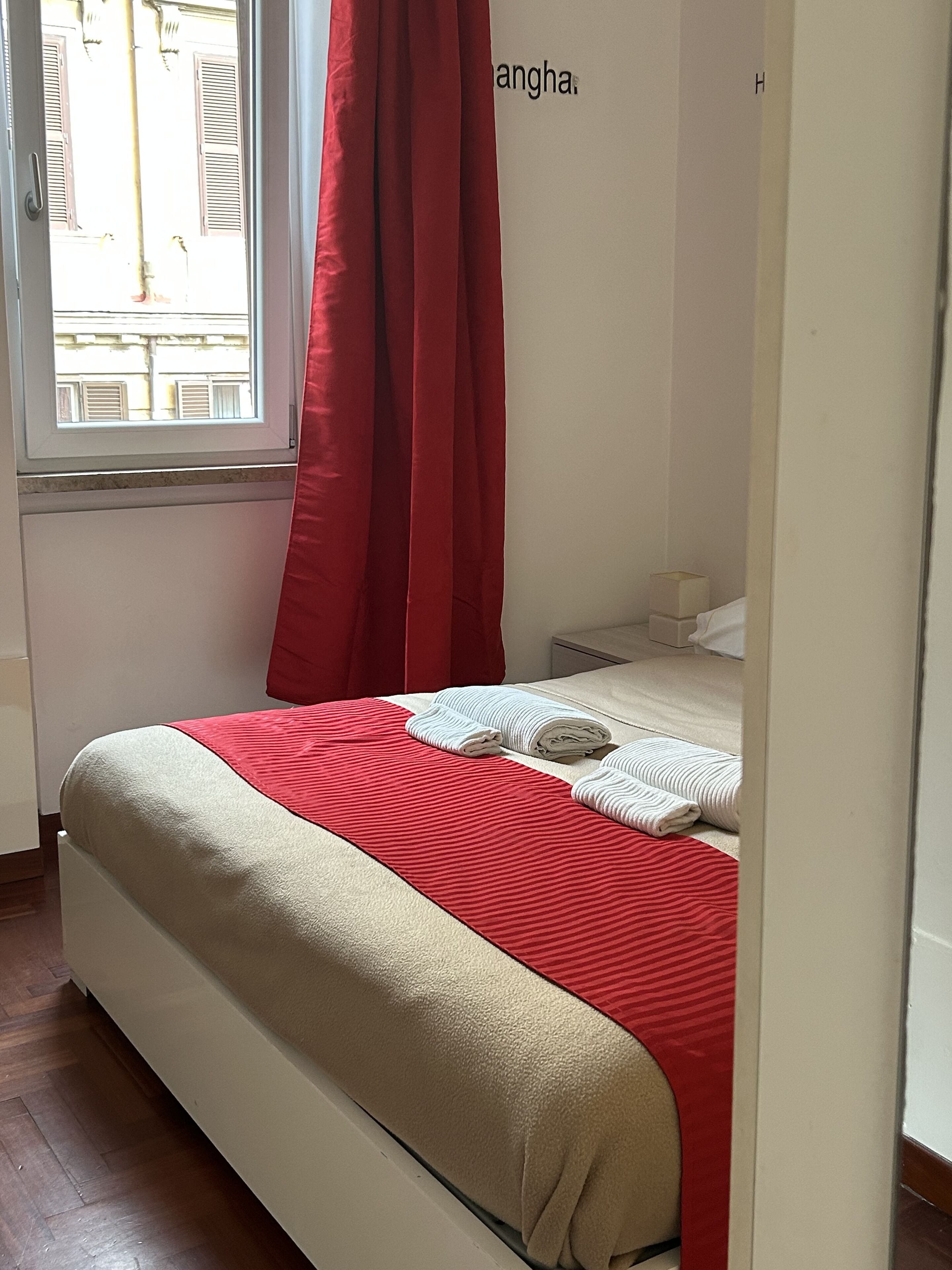 A hotel room with a bed, red curtains, a window with shutters, and a wall with the word "Shanghai".