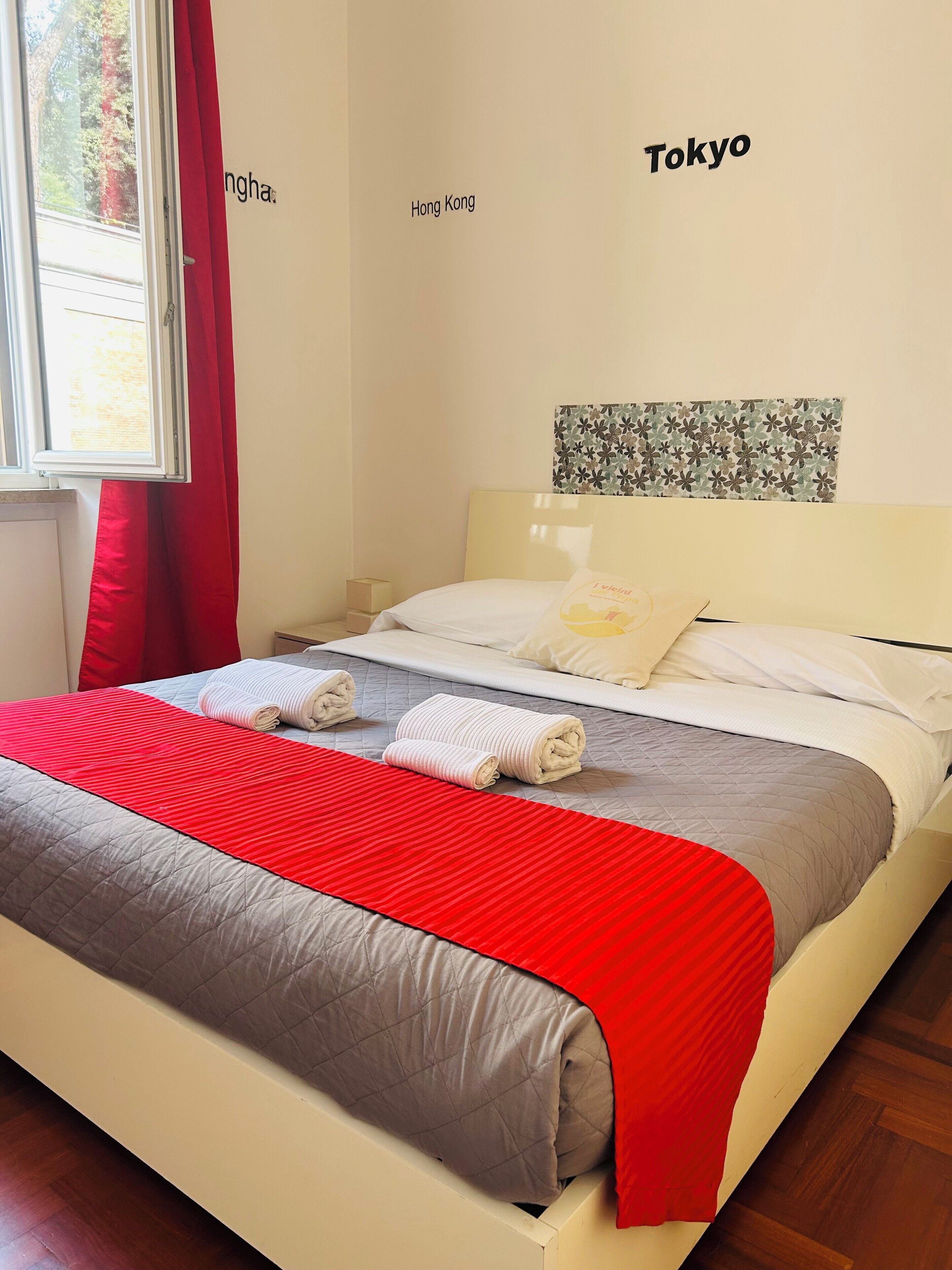 Een net opgemaakt bed met een rood-grijs dekbed, witte kussens en opgerolde handdoeken. De kamer heeft een raam met rode gordijnen, een muur met de tekst 'Hong Kong' en 'Tokyo' erop, en een bloemistisch kunstwerk boven het bed.