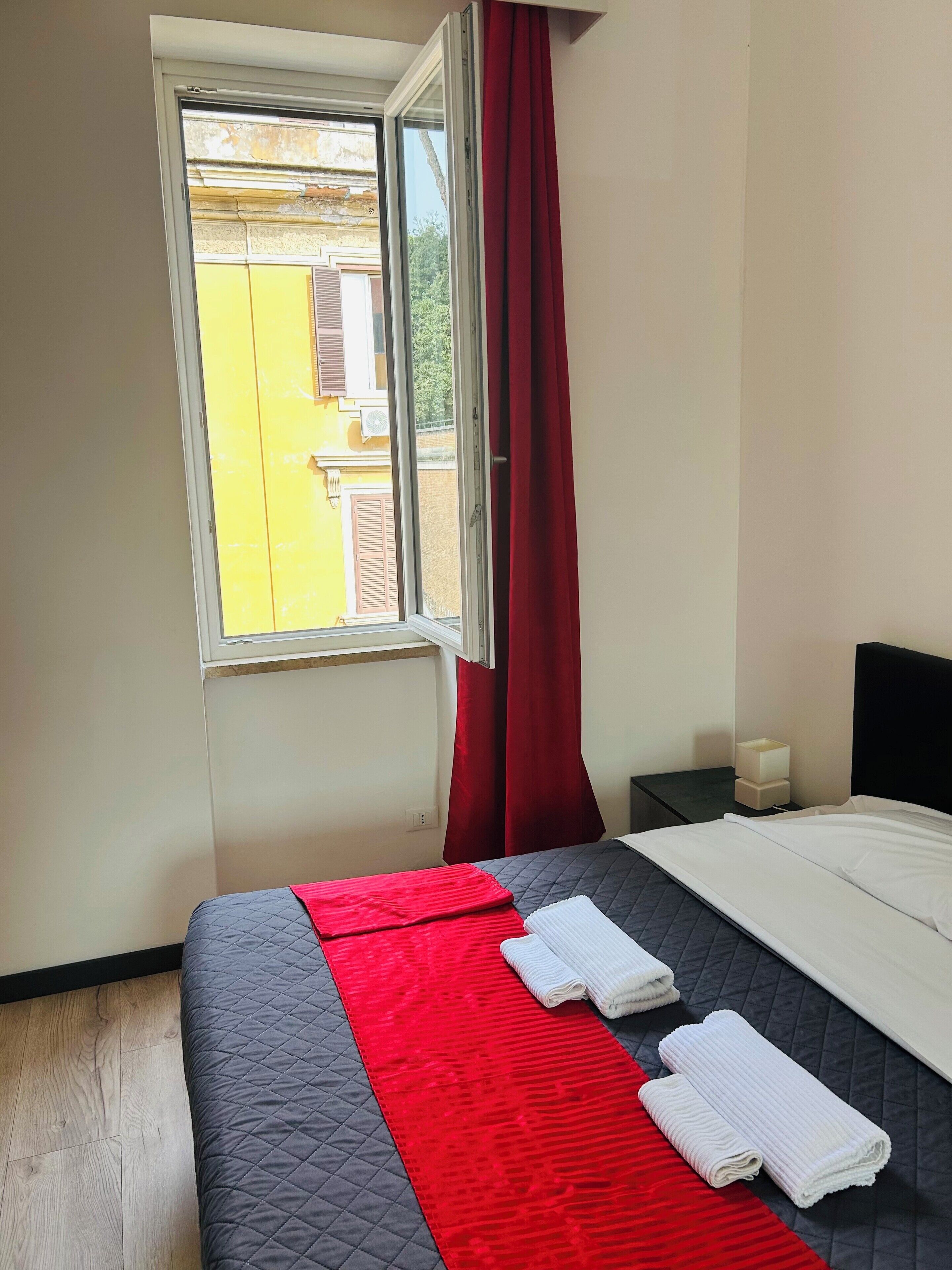 A hotel room with a bed, a window with red curtains, a bedside table with a lamp, and a view of buildings outside.