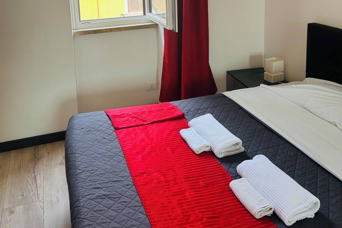 A hotel room with a bed, a window with red curtains, a bedside table with a lamp, and a view of buildings outside.