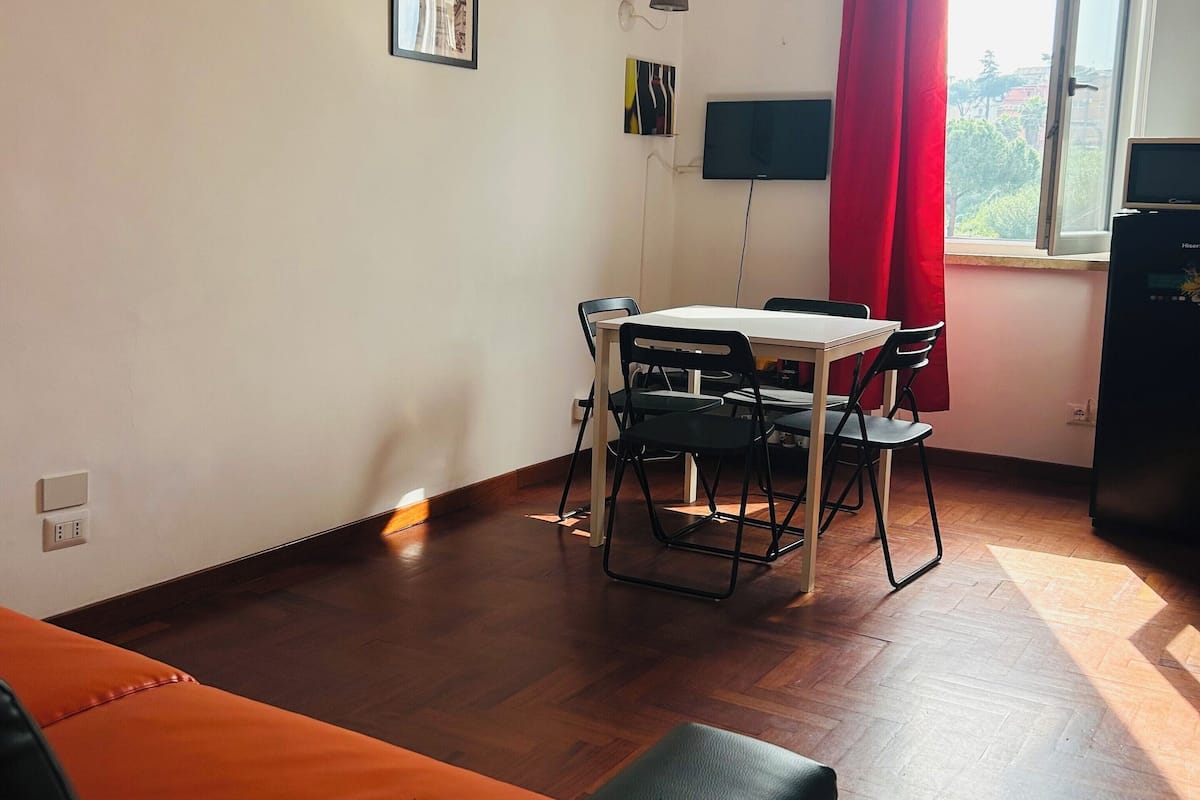 A room with a dining table and chairs, a sofa, a TV, and a window with red curtains.