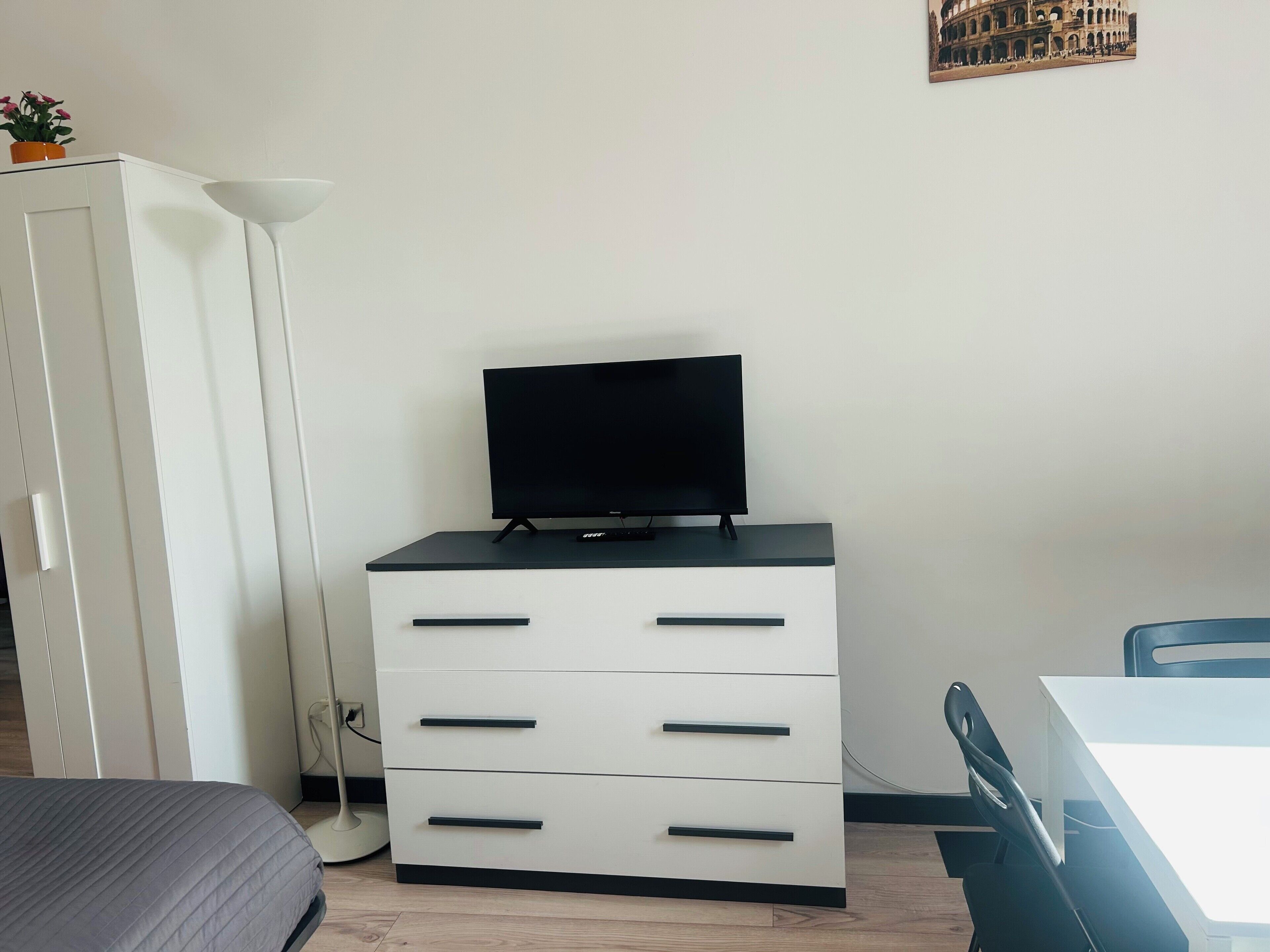 A bedroom with a bed, a TV on a dresser, a lamp, and a picture of the Colosseum on the wall.