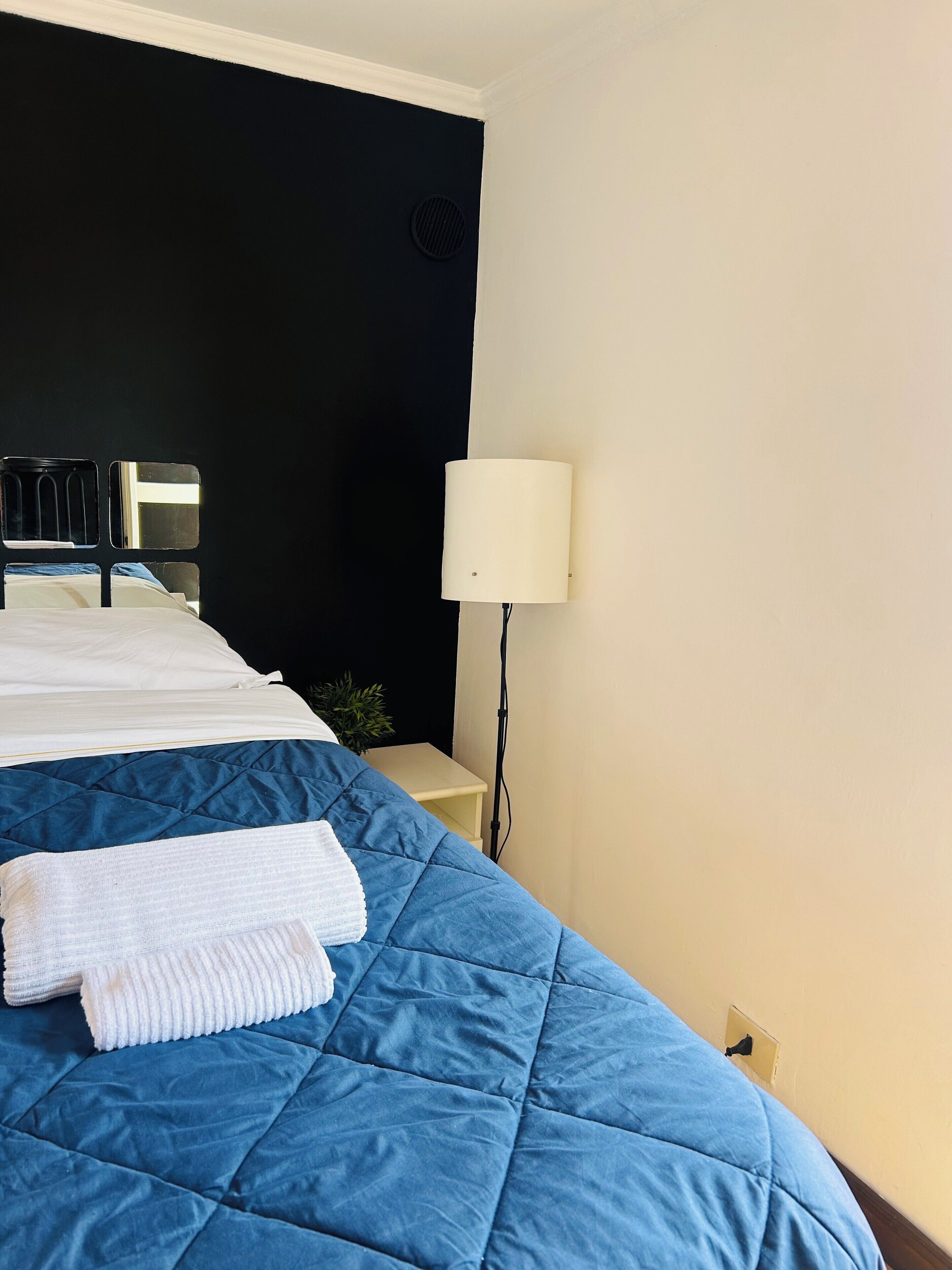A neatly made bed with a blue quilt and a white towel, a bedside lamp, and a wall-mounted mirror.