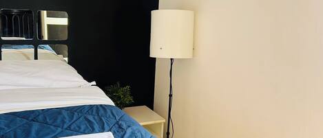 Cama arrumada com um edredom azul e uma toalha branca, luminária de cabeceira e espelho fixado na parede.