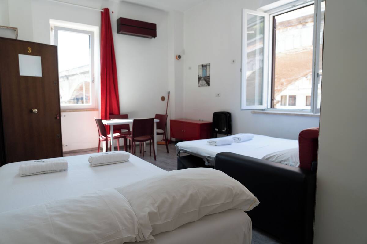 A hotel room with two beds, a desk, a chair, a red curtain, and a window with a view.