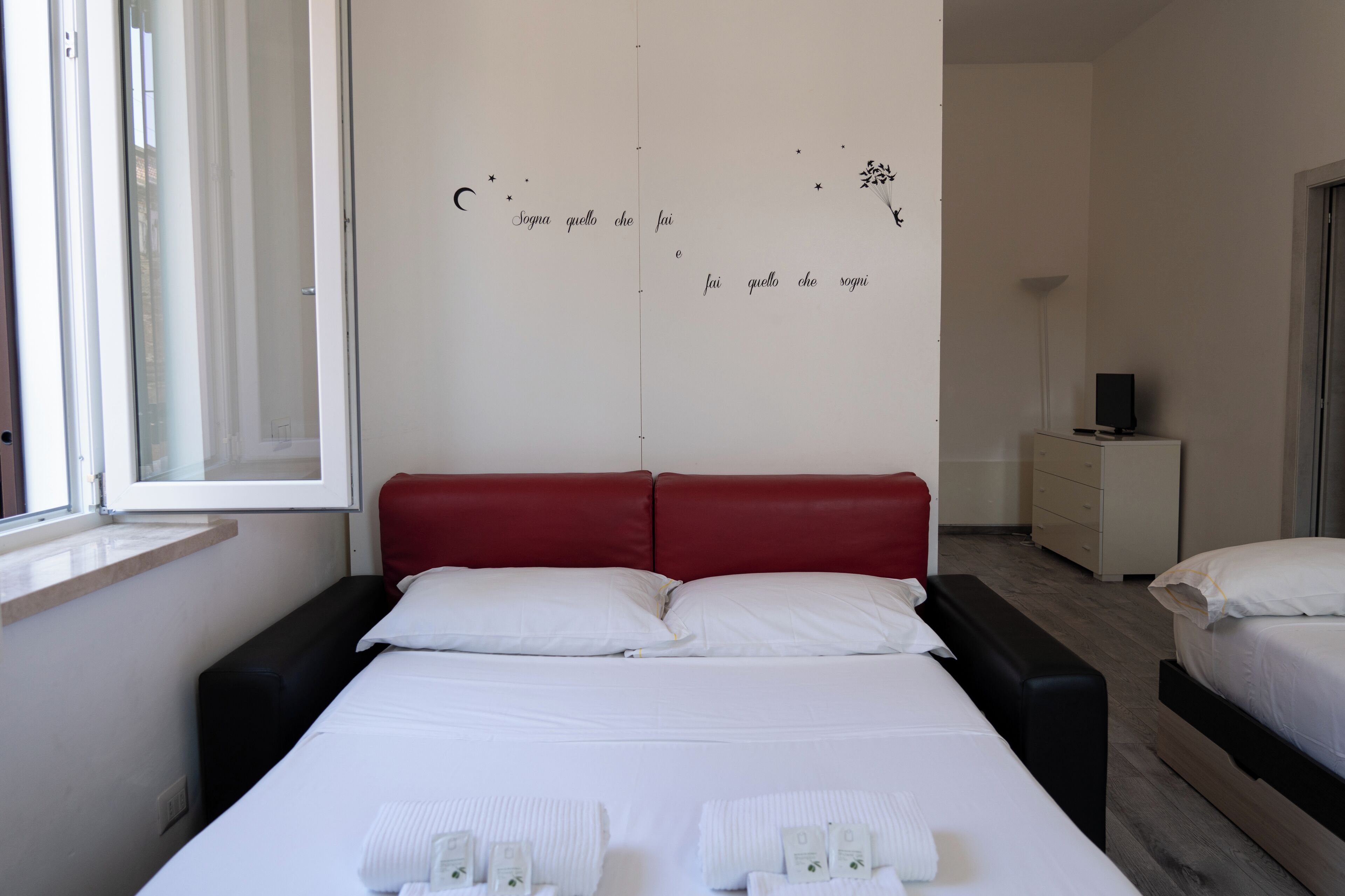 Une chambre d’hôtel avec un grand lit, une tête de lit rouge, du linge de lit blanc et un mur orné d’une inscription décorative.