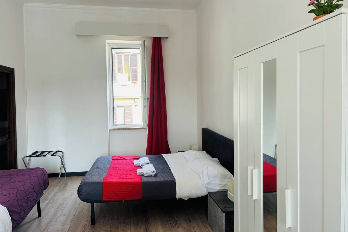 A bedroom with a bed, a window with red curtains, a wardrobe, and a small table.